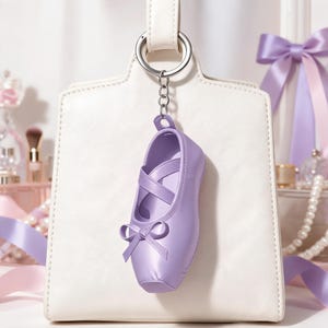 Puede incluir: Un llavero de zapatilla de ballet color lavanda con acabado liso, tiras cruzadas en la parte superior y un peque&ntilde;o lazo decorativo en la punta. El zapato est&aacute; unido a una anilla de metal plateado mediante una cadena corta.