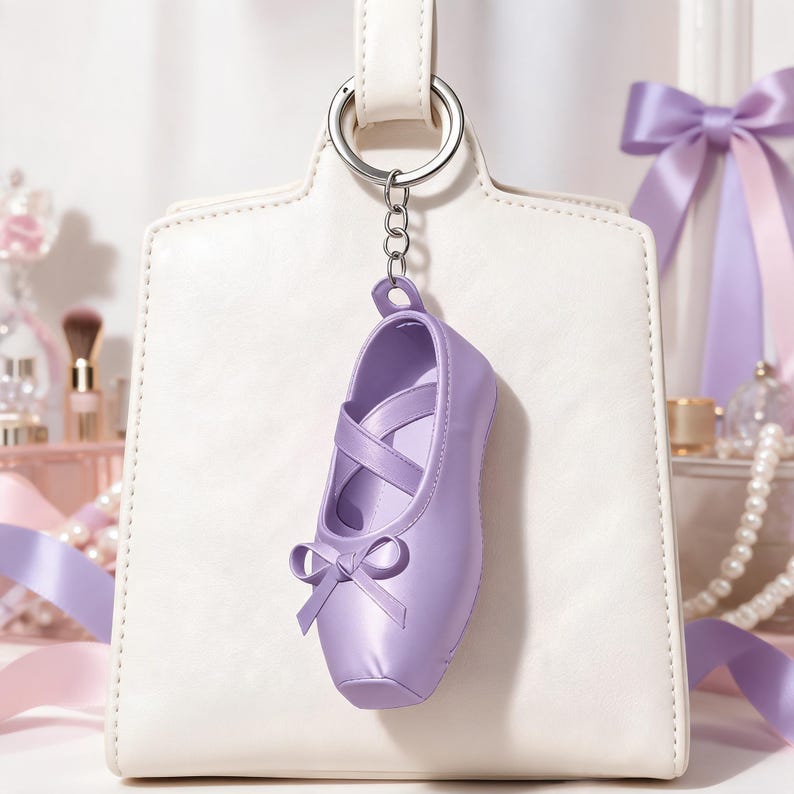 Puede incluir: Un llavero de zapatilla de ballet color lavanda con acabado liso, tiras cruzadas en la parte superior y un peque&ntilde;o lazo decorativo en la punta. El zapato est&aacute; unido a una anilla de metal plateado mediante una cadena corta.