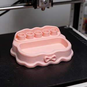 May include: A light pink 3D printed organiser tray featuring a wavy edge, a central rectangular compartment, and five cylindrical slots along the back. The front of the tray is decorated with a small, centred bow detail and horizontal layer lines.