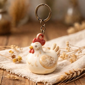 May include: A keyring featuring a small, cream-coloured chicken figurine with a red comb and wattle, a yellow beak, and light brown wing details. The chicken is attached to a circular, antique-bronze coloured metal ring by a short chain link.