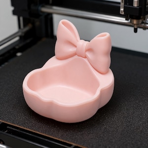 Puede incluir: Un cuenco de color rosa claro, liso y con forma de cuenco, que presenta un detalle de lazo moldeado prominente en el borde posterior. El objeto tiene una base redondeada y festoneada y un interior cóncavo, con un color pastel uniforme.