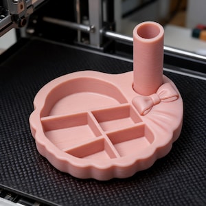 Op de afbeelding: Een lichtroze 3D-geprinte organizer-tray met een cilindrische pennenhouder, een kleine decoratieve strik en vier verdeelde vakken. De tray heeft een geschulpte rand en is gemaakt van een mat, getextureerd kunststof materiaal.