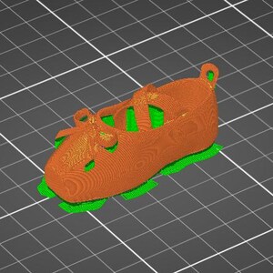 Puede incluir: Un modelo digital 3D de un zapato con un exterior naranja texturizado, un detalle de lazo en la parte delantera y una correa en la parte superior. El zapato est&aacute; colocado sobre una base de color verde brillante sobre un fondo de cuadr&iacute;cula gris oscuro.