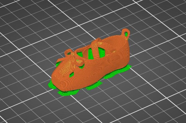 Puede incluir: Un modelo digital 3D de un zapato con un exterior naranja texturizado, un detalle de lazo en la parte delantera y una correa en la parte superior. El zapato est&aacute; colocado sobre una base de color verde brillante sobre un fondo de cuadr&iacute;cula gris oscuro.