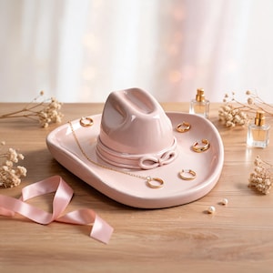Peut inclure: Un vide-poche en céramique rose pâle en forme de chapeau de cowboy avec une finition lisse et brillante. Le plat est orné d'un collier à chaîne dorée délicat et de plusieurs petites créoles dorées posées sur le bord, avec un ruban rose.