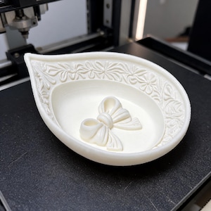 May include: A teardrop-shaped off-white dish featuring a raised floral pattern along the inner rim and a three-dimensional bow centered at the base. The object has a matte finish and is constructed with visible horizontal 3D printing layers.