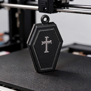 Puede incluir: Un colgante negro con forma de ataúd que presenta un emblema de cruz de color plateado centrado en la parte frontal. El colgante tiene un acabado texturizado y un lazo circular en la parte superior para su sujeción, con un diseño geométrico estructurado.