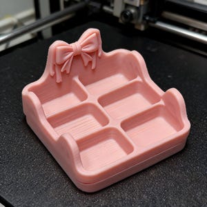 May include: A light pink 3D printed organiser tray featuring four rectangular compartments and a decorative bow detail on the back wall. The tray has a smooth, matte finish with visible horizontal layer lines from the printing process.
