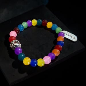 May include: A multi-colored beaded bracelet featuring smooth, round, translucent beads in shades of red, orange, yellow, green, blue, purple, and pink. The bracelet includes a silver-toned metal spacer bead and a rectangular silver charm with the word Create.