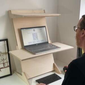 May include: A light-colored wooden standing desk with a laptop on the main surface and a shelf above. A black keyboard sits on the desk. The laptop displays a website. A person is using the desk.