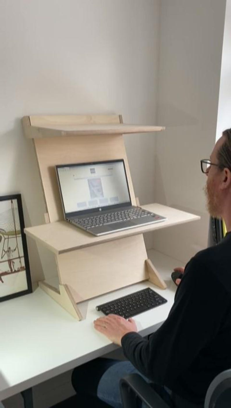 May include: A light-colored wooden standing desk with a laptop on the main surface and a shelf above. A black keyboard sits on the desk. The laptop displays a website. A person is using the desk.
