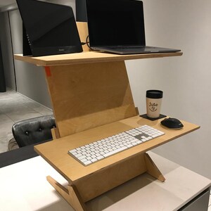 May include: A two-tiered, light-colored wooden standing desk with a laptop, monitor, keyboard, mouse, and a coffee cup. The desk has a unique angled design and is set up for a workspace.