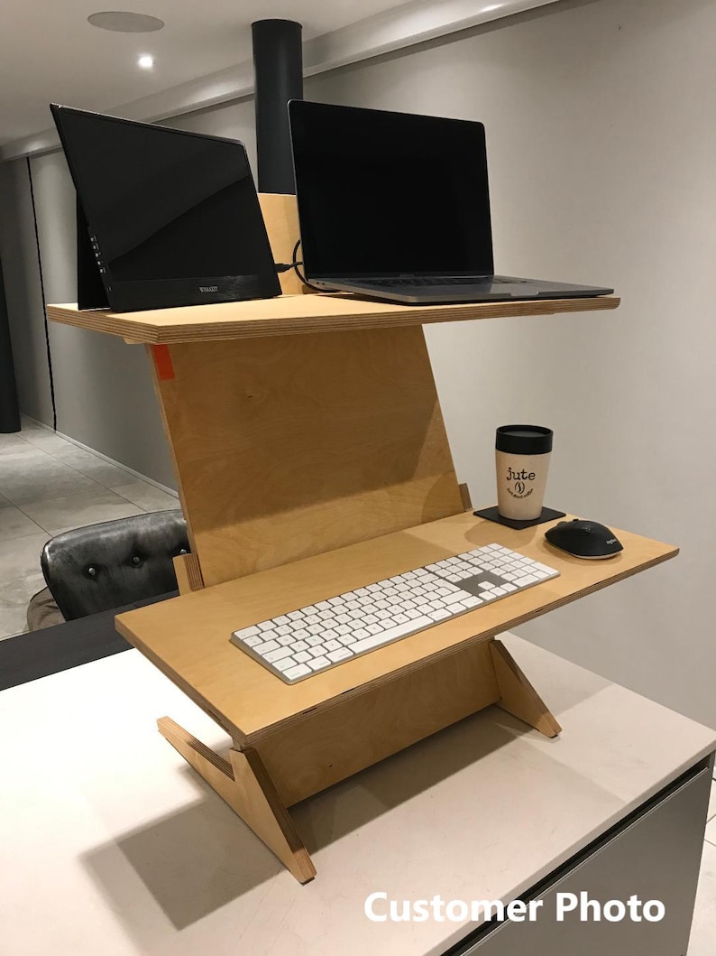 May include: A two-tiered, light-colored wooden standing desk with a laptop, monitor, keyboard, mouse, and a coffee cup. The desk has a unique angled design and is set up for a workspace.