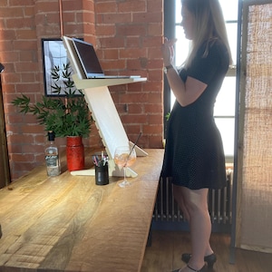 May include: A light-colored wooden laptop stand with a laptop on top. The stand is on a large wooden table. A glass of orange-colored drink sits on the table. A woman in a black dress stands near the table.