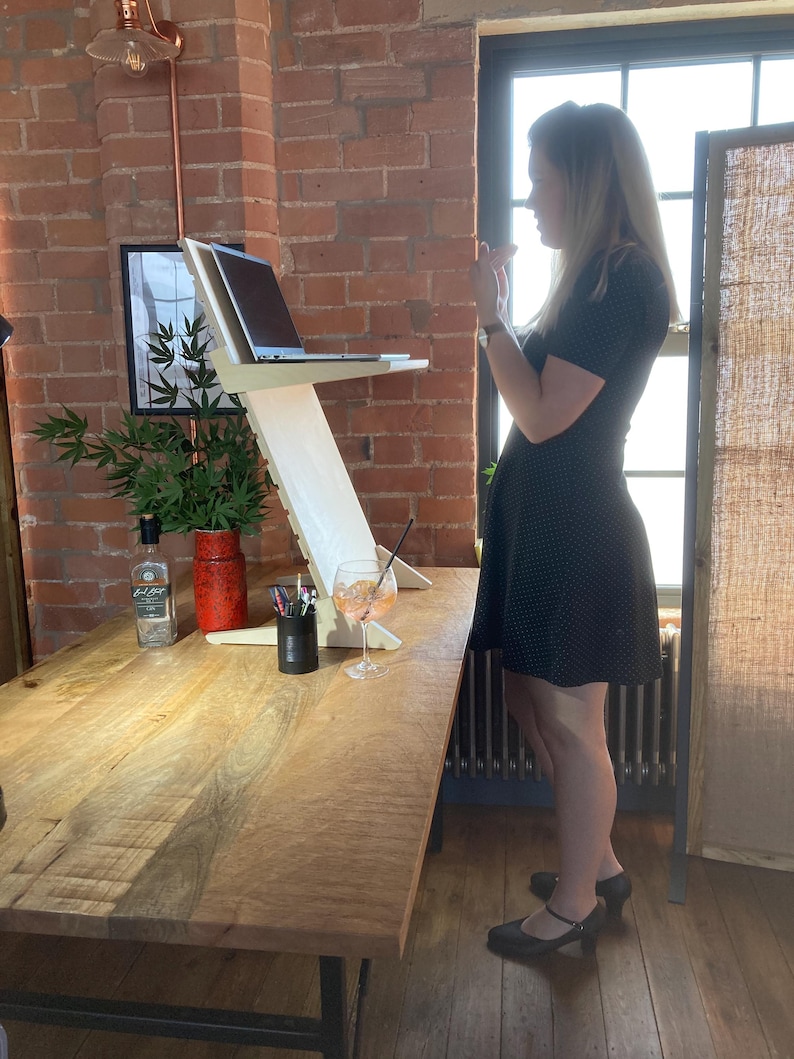 May include: A light-colored wooden laptop stand with a laptop on top. The stand is on a large wooden table. A glass of orange-colored drink sits on the table. A woman in a black dress stands near the table.