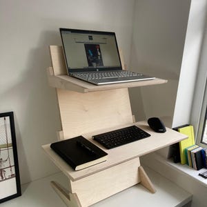 May include: A light-colored wooden standing desk with two tiers. The top tier holds a laptop, and the lower tier has a keyboard, mouse, and a black notebook. The desk is positioned in a corner near a window.
