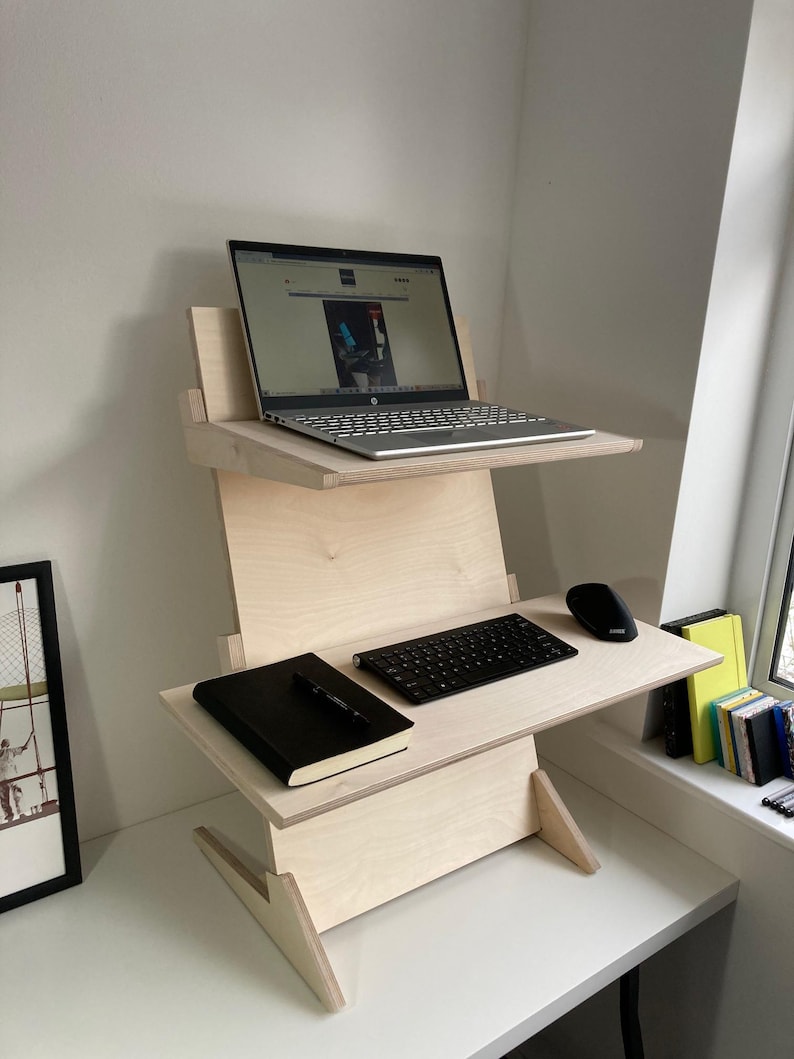 May include: A light-colored wooden standing desk with two tiers. The top tier holds a laptop, and the lower tier has a keyboard, mouse, and a black notebook. The desk is positioned in a corner near a window.