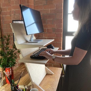 May include: A wooden standing desk with a computer monitor and keyboard. The desk has multiple levels and is made of light-colored wood. A person is using the keyboard. The desk is on a wooden table. A mug and art supplies are on the table.