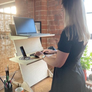 May include: A light-colored wooden standing desk with two tiers, holding a laptop and a smartphone. The desk is positioned in front of a brick wall, with a mug and art supplies visible on the desk. A person is using the desk.