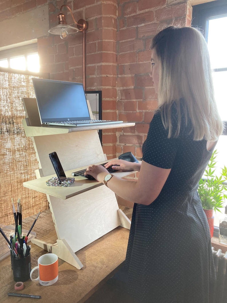 May include: A light-colored wooden standing desk with two tiers, holding a laptop and a smartphone. The desk is positioned in front of a brick wall, with a mug and art supplies visible on the desk. A person is using the desk.