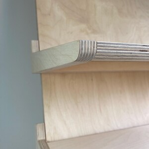 May include: Close-up of a light-colored wooden shelf with a curved edge. The shelf is made of layered wood, with visible grain patterns. The design features a minimalist aesthetic, showcasing the natural wood tones and textures.
