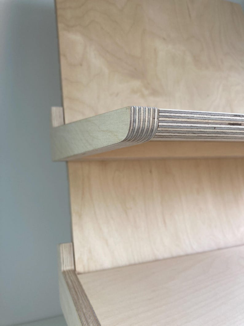May include: Close-up of a light-colored wooden shelf with a curved edge. The shelf is made of layered wood, with visible grain patterns. The design features a minimalist aesthetic, showcasing the natural wood tones and textures.