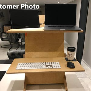 May include: A two-tiered, light-colored wooden standing desk with a monitor and laptop on the top tier. The bottom tier holds a keyboard and mouse. A mug and coaster sit on the side. The text "Customer Photo" is in the top left corner.