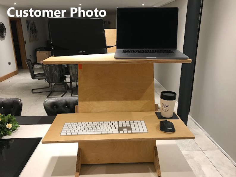 May include: A two-tiered, light-colored wooden standing desk with a monitor and laptop on the top tier. The bottom tier holds a keyboard and mouse. A mug and coaster sit on the side. The text "Customer Photo" is in the top left corner.