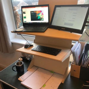 May include: A wooden standing desk with two tiers, holding a laptop and a large monitor. A black keyboard, mouse, and a mug are on the desk. The desk also has a notepad and papers. The text "Customer Photo" is visible.