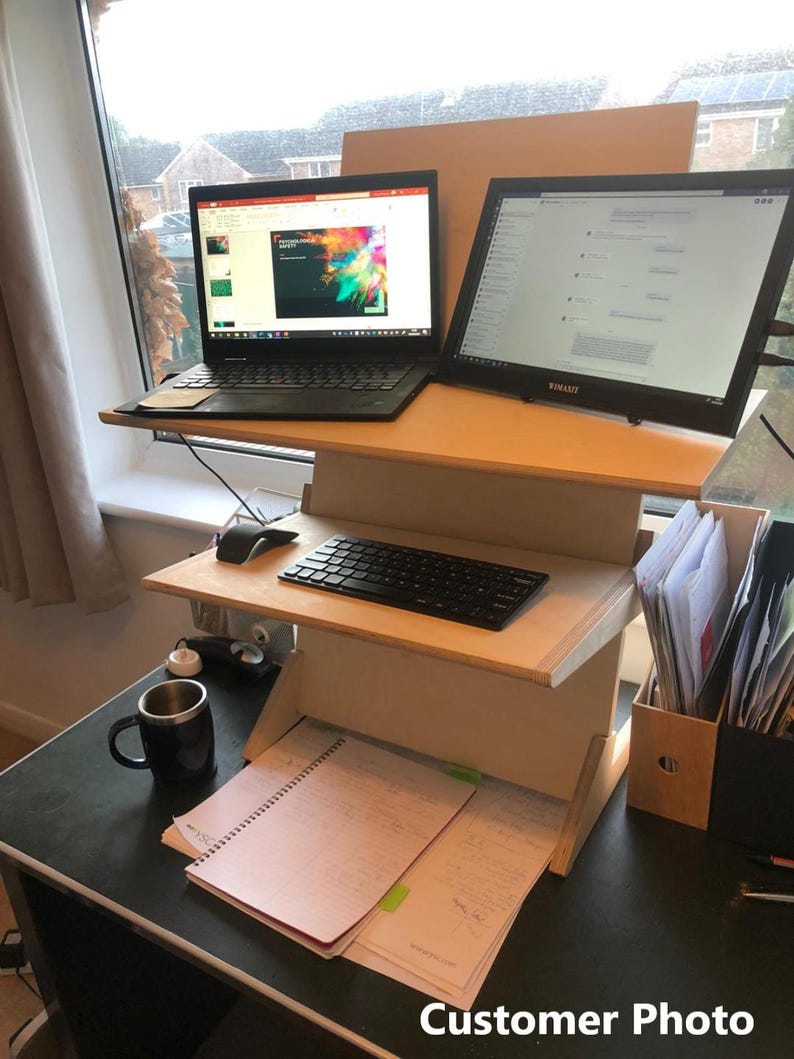 May include: A wooden standing desk with two tiers, holding a laptop and a large monitor. A black keyboard, mouse, and a mug are on the desk. The desk also has a notepad and papers. The text "Customer Photo" is visible.