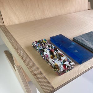 May include: A wooden desk with a light finish, featuring rectangular blocks of various colors and textures. One block is a mix of colors, another is blue, and a third is gray. The desk has a simple, modern design.