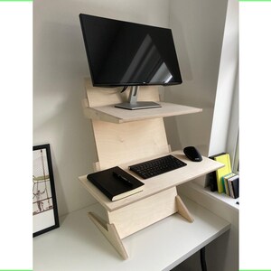 May include: A light-colored wooden standing desk with two shelves. A black computer monitor sits on the top shelf. The lower shelf holds a keyboard, mouse, and a black notebook. The desk is on a white surface.