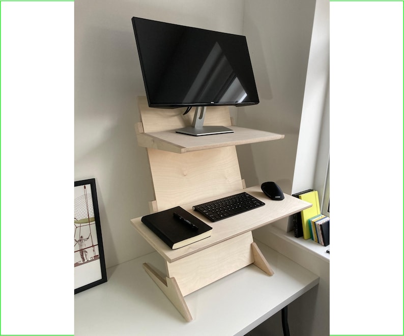 May include: A light-colored wooden standing desk with two shelves. A black computer monitor sits on the top shelf. The lower shelf holds a keyboard, mouse, and a black notebook. The desk is on a white surface.