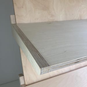 May include: Close-up of a light-colored wooden shelf with a layered edge. The shelf is part of a larger wooden structure, possibly a bookcase or shelving unit. The wood grain is visible, and the overall design is minimalist.
