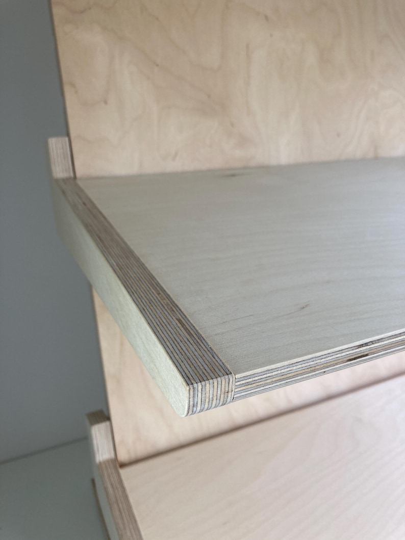 May include: Close-up of a light-colored wooden shelf with a layered edge. The shelf is part of a larger wooden structure, possibly a bookcase or shelving unit. The wood grain is visible, and the overall design is minimalist.