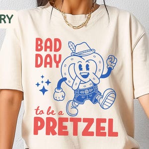 May include: An ivory short-sleeve t-shirt featuring a graphic print in red and blue. The design includes the text "BAD DAY to be a PRETZEL" alongside a cartoon illustration of a pretzel wearing a hat and shorts, accompanied by small star icons.