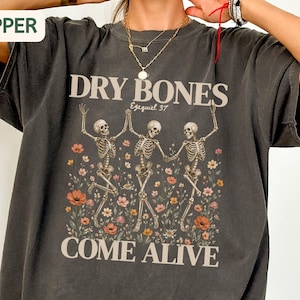 May include: A charcoal gray short-sleeve t-shirt featuring a graphic of three skeletons dancing among colorful wildflowers. The text reads DRY BONES, Ezequiel 37, and COME ALIVE in a cream-colored font. The shirt has a crew neckline and a relaxed fit.
