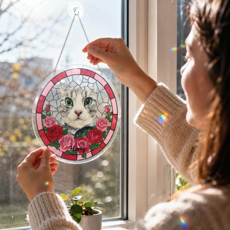 Person hanging a round acrylic puzzle suncatcher on a sunny window using a suction hook and silver chain. The custom pet portrait design features a gray cat with green eyes, pink and red roses, a stained glass style border, and the name “Harry,” with rainbow light reflections shining across the window and sweater.
