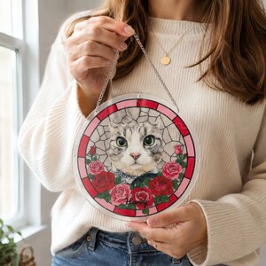 Person holding a round acrylic puzzle suncatcher featuring a realistic cat face with pink and red roses, surrounded by a pink and red decorative border, hanging by a stainless steel chain, with the person wearing a white sweater and blue jeans, photographed near a window.