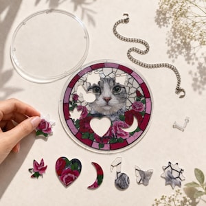 DIY acrylic puzzle suncatcher assembly scene on a light tabletop, featuring a custom gray cat portrait with pink and red roses in a round clear frame. Irregular puzzle pieces shaped like a star, heart, moon, butterfly, flower, and music note are placed around it, with a stainless steel hanging chain, screws, and clear acrylic cover nearby.