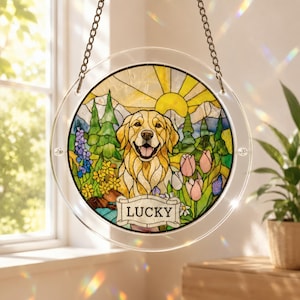 Round acrylic puzzle suncatcher hanging by a stainless steel chain in front of a sunny window, featuring a happy golden retriever surrounded by colorful flowers and green trees, with a yellow sun rising in the background. The suncatcher has a clear border and a nameplate at the bottom reading ‘LUCKY,’ with soft rainbow light reflections on the wall and windowsill nearby.