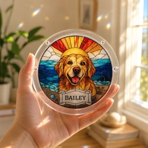 Hand holding a small round acrylic puzzle suncatcher near a sunny window, featuring a golden retriever portrait with a colorful beach sunset background, blue ocean waves, orange sun rays, and a custom nameplate reading “BAILEY.” The clear acrylic frame shows screw details and light-catching rainbow reflections in the room.