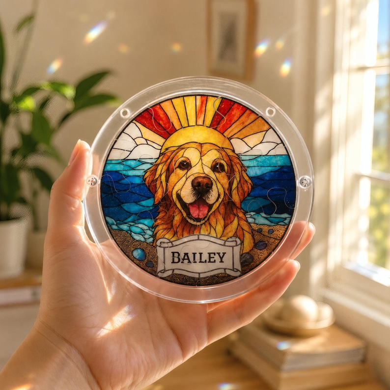 Hand holding a small round acrylic puzzle suncatcher near a sunny window, featuring a golden retriever portrait with a colorful beach sunset background, blue ocean waves, orange sun rays, and a custom nameplate reading “BAILEY.” The clear acrylic frame shows screw details and light-catching rainbow reflections in the room.