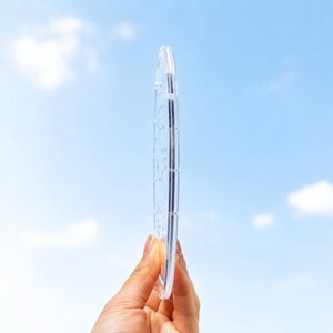 Side view of a round acrylic puzzle suncatcher held by hand against a bright blue sky, showing the clear layered acrylic construction and slim transparent edge. The image highlights the thickness, smooth finish, and durable material of the custom hanging puzzle ornament.