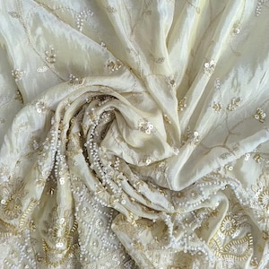 Puede incluir: Una tela de color crema con intrincados bordados de hilo metálico dorado, lentejuelas transparentes y pequeñas cuentas blancas. El material está drapeado en pliegues suaves, mostrando un patrón floral y abalorios densos en los bordes.