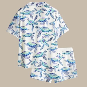 May include: White pajama set featuring a pattern of blue, green, and purple sea turtles. The set includes a short-sleeved button-up shirt and matching shorts. The edges of the shirt and shorts are trimmed with a thin blue border.