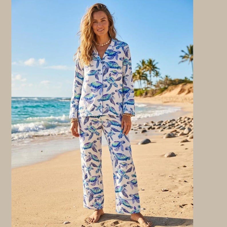 May include: White pajama set with a blue and green sea turtle pattern. The long-sleeved top has a button-down front and the pants are straight-legged. The pajamas are worn on a sandy beach with the ocean in the background.