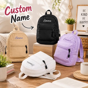 Könnte beinhalten: Vier kleine Rucksäcke in Schwarz, Beige, Weiß und Helllila, jeweils mit dem Namen Emma in Schreibschrift auf der Vordertasche bestickt. Jeder Rucksack verfügt über ein Hauptfach mit Reißverschluss, eine kleinere Vordertasche und Träger.