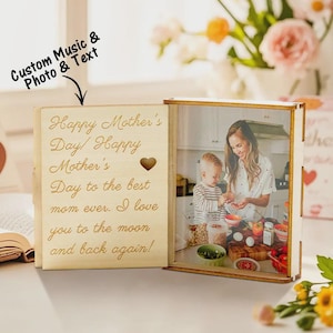 Puede incluir: Una caja de fotos de madera con una foto de una madre y un niño. La caja está abierta, revelando una foto y un texto grabado que dice "Feliz Día de la Madre". La caja está rodeada de flores y otros elementos decorativos.