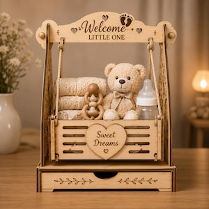 Pode incluir: Um conjunto de presente para bebé em madeira em forma de baloiço com um urso de peluche claro, um guizo de madeira, um biberão e tecido dobrado. A estrutura tem gravado "Welcome LITTLE ONE" e o cesto um coração com "Sweet Dreams".
